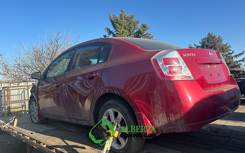 Red Nissan Sentra with side damage loaded on a tow trailer under clear blue sky at Alberta Cash for Cars