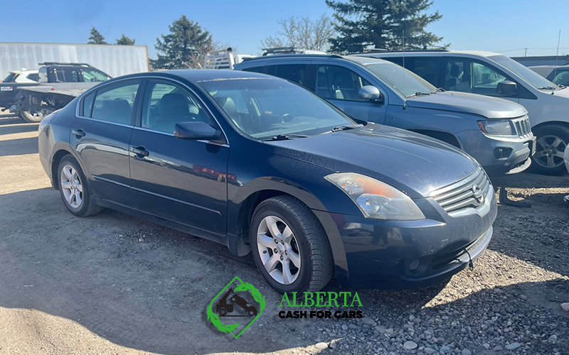 Dark blue Nissan Altima sedan parked in a sunny dirt junkyard, scrap cars visible.