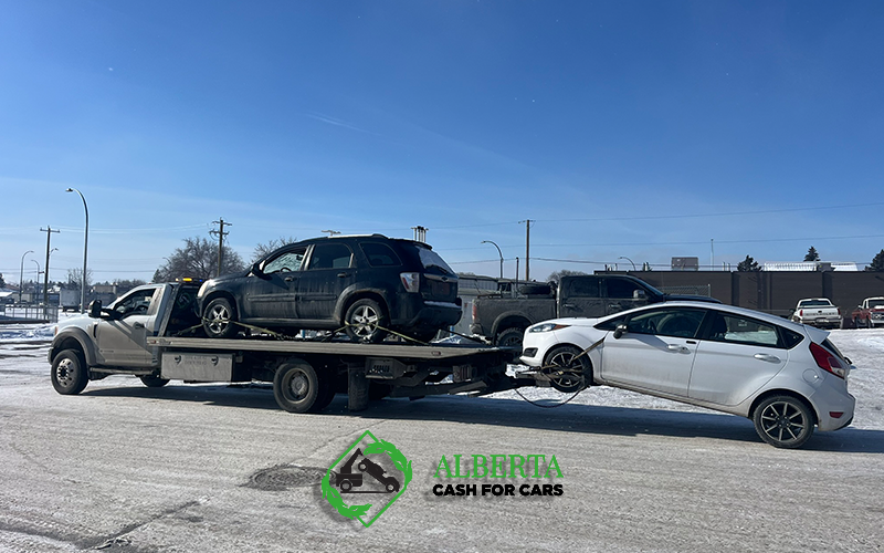 Tow truck carrying black SUV and white Ford Fiesta in Alberta snow; "Cash for Cars" logo.