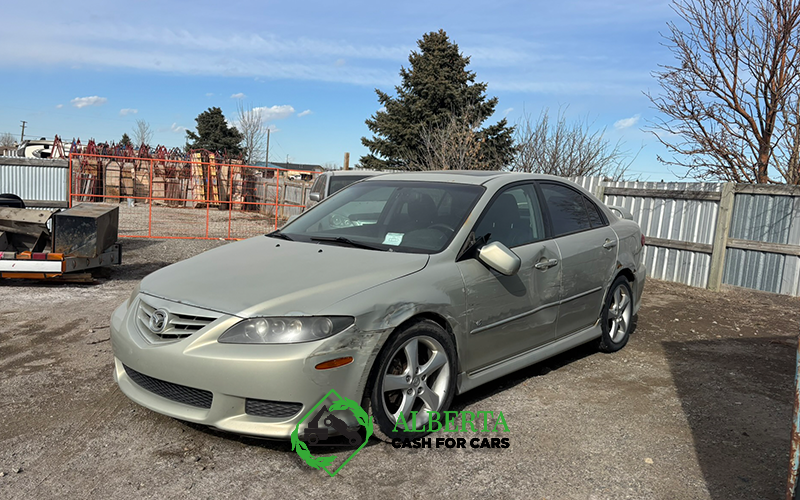 Silver Mazda 6 sedan with damaged fender in a salvage yard under a bright sky. Text: Alberta Cash for Cars.