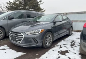 Grey Hyundai Elantra sedan parked in a snowy, muddy lot next to a minivan and a metal fence.