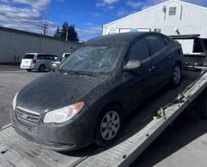Dirty dark grey Hyundai Elantra sedan on a flatbed tow truck, parked outside "I&D AUTO" garage under a blue sky.