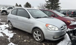 Silver Hyundai Accent hatchback parked in a muddy, snowy salvage yard, surrounded by other damaged cars.