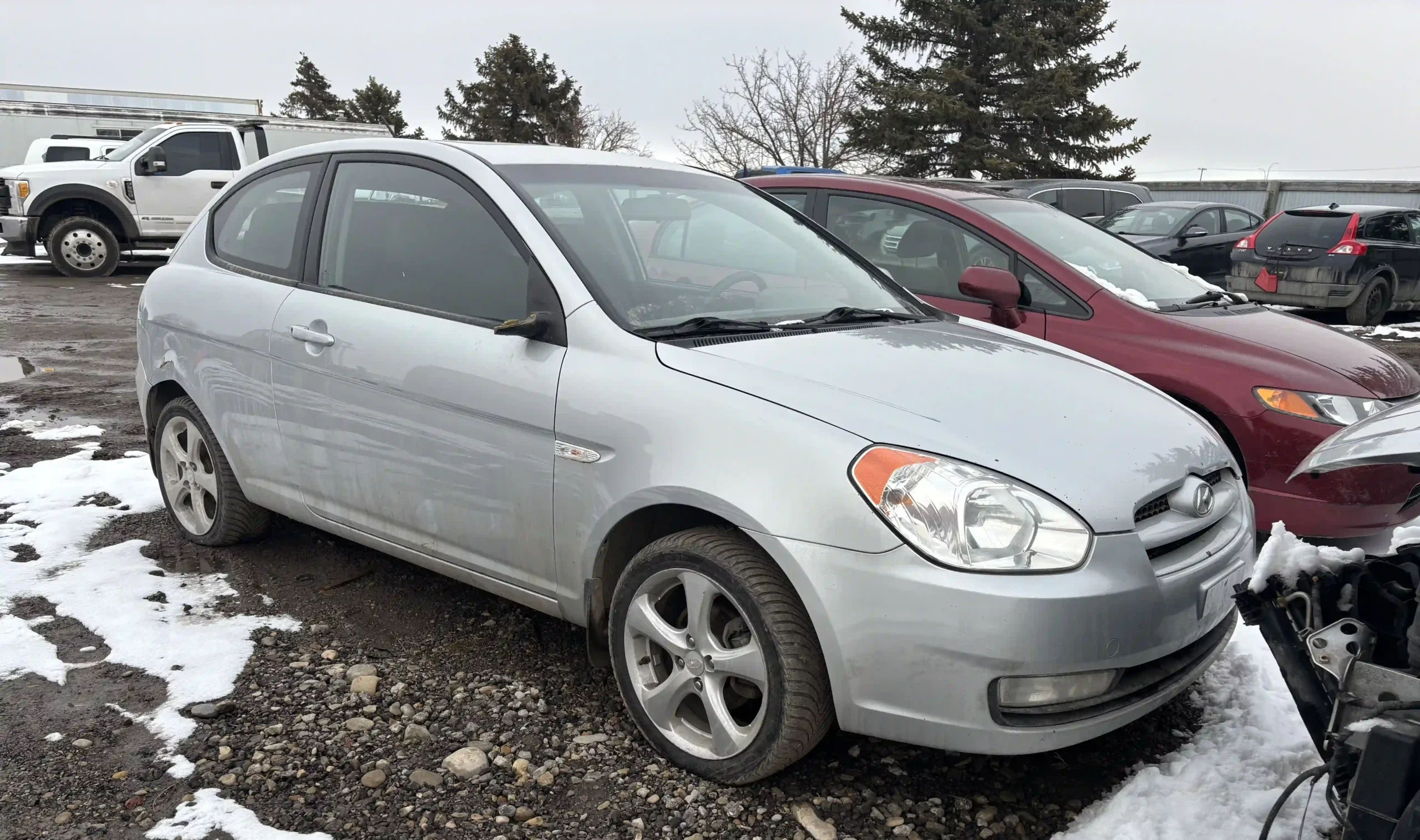 Silver Hyundai Accent hatchback parked in a muddy, snowy salvage yard, surrounded by other damaged cars.