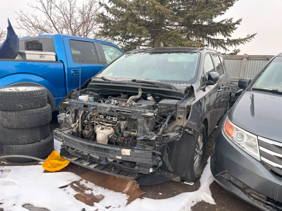 Wrecked dark grey Toyota Sienna with a completely removed front bumper, engine visible, in a snowy salvage lot.