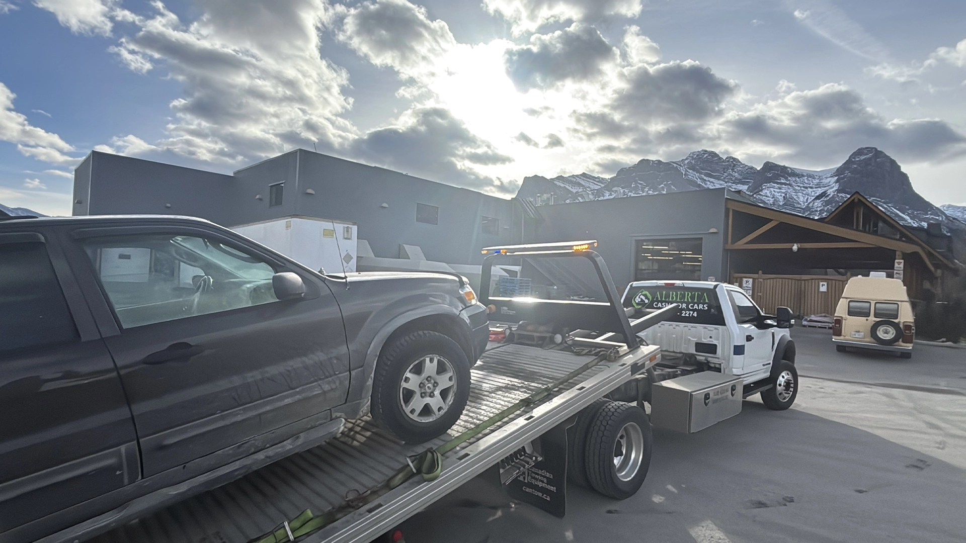 A white flatbed tow truck loading a dark gray SUV in a parking lot, with snow-capped mountains and a commercial building in the background under a cloudy, bright sky.