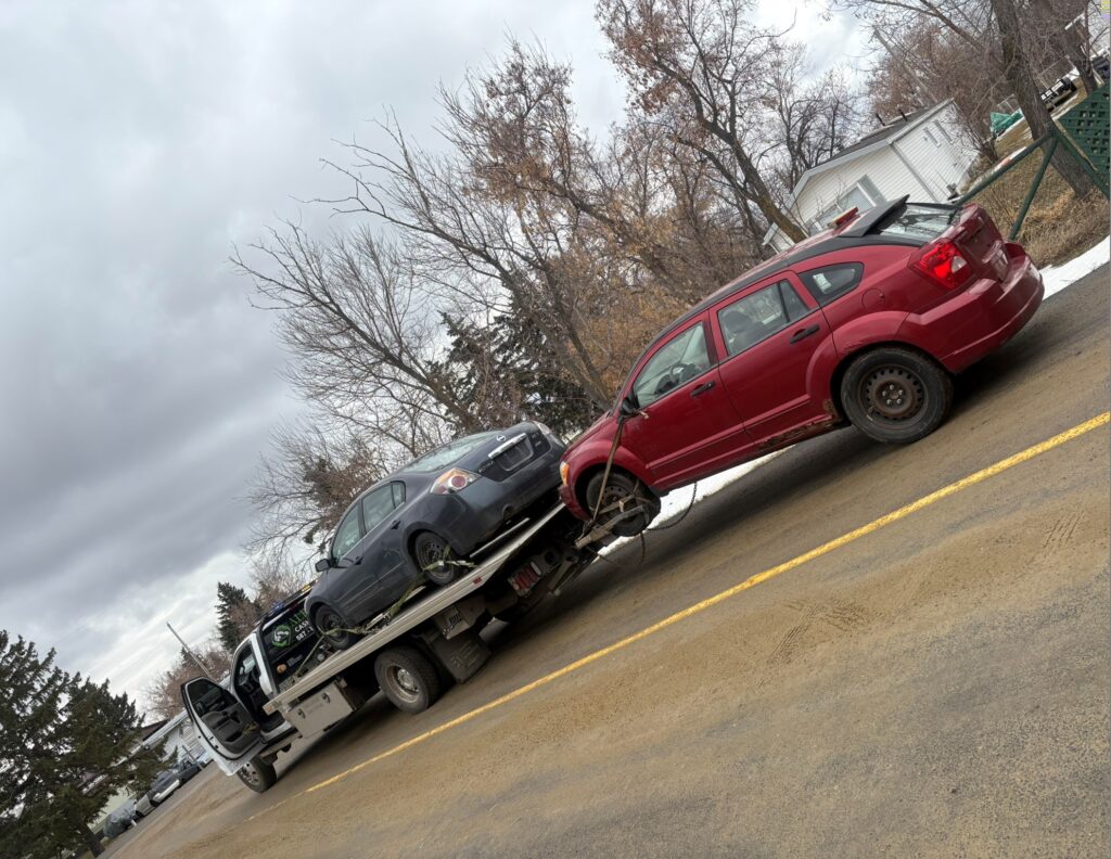 Red and dark gray cars on a flatbed tow truck in Diamond Valley, Alberta on a cloudy day.