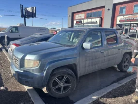 Blue Honda Ridgeline pickup truck parked in a lot in front of commercial buildings on a sunny day.