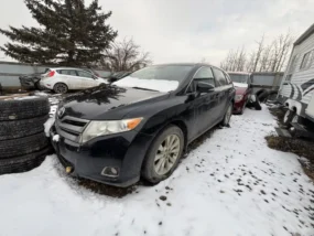Black Toyota Venza parked in a snowy junkyard with other vehicles and stacked tires.