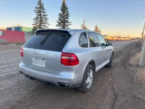 Silver Porsche Cayenne SUV on a dirt road at sunset with shipping containers in the distance.