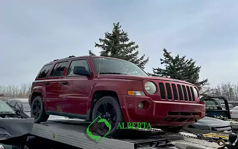 Red Jeep SUV on flatbed tow truck, Alberta Cash For Cars sign 587-844-2274, clear blue sky, evergreen trees, car lot surroundings.