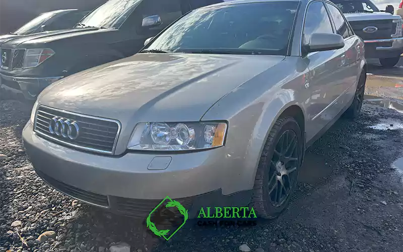  Silver Audi sedan parked on gravel lot, sunny weather, cloudy sky, surrounded by Nissan Frontier and Ford trucks, wet ground.