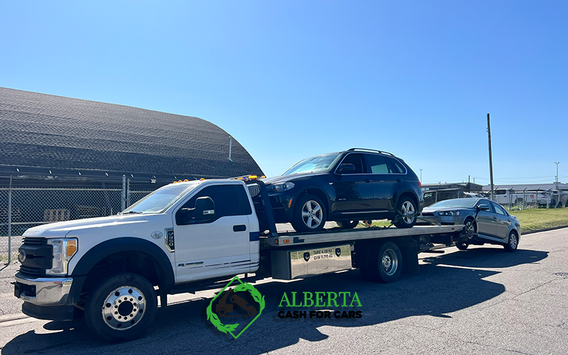 Large white tow truck carrying a dark SUV and a gray sedan on a flatbed; blue sky; "ALBERTA CASH FOR CARS" logo.