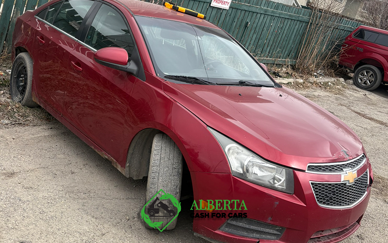 Damaged red Chevrolet sedan with a flat tire on a dirt lot, visible front-end and missing headlight.
