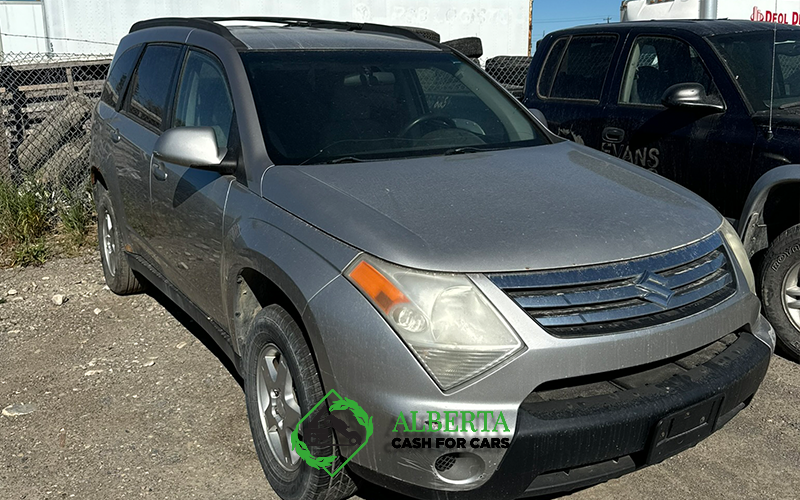 Silver Suzuki XL7 SUV parked in a salvage lot on a sunny day with another truck in the background.