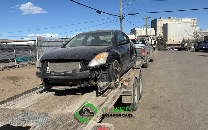 damaged black sedan on trailer in sunny industrial street, Alberta Cash for Cars towing visible