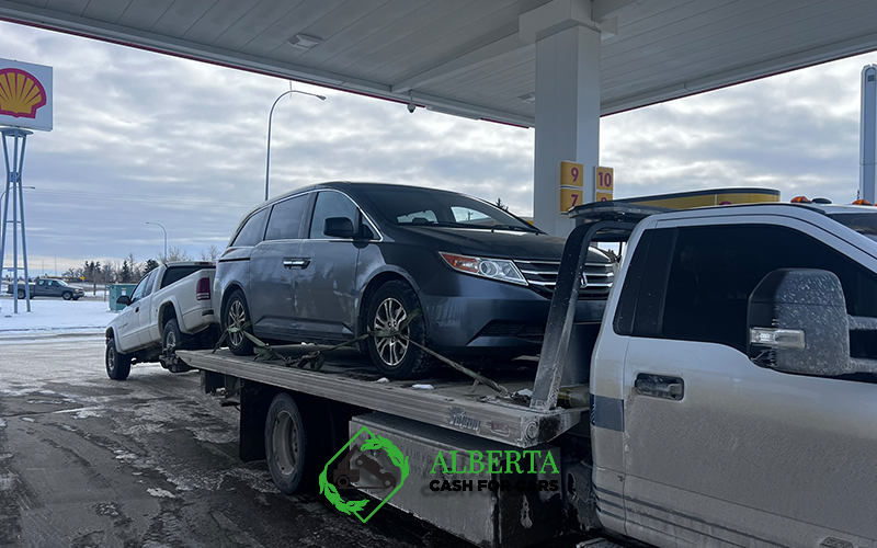 Tow truck carrying a grey minivan and white pickup near Shell gas station on snowy Alberta roadside