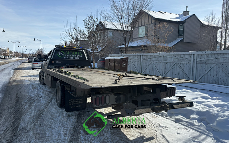 Alberta Cash for Cars flatbed tow truck parked on a snowy residential street with houses and winter daylight.