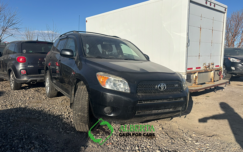 Black Toyota RAV4 V6 parked on a gravel lot next to a white trailer, representing a vehicle possibly designated for scrap or engine service