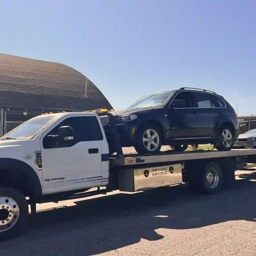 White flatbed tow truck carrying a dark SUV and towing a silver sedan on a sunny industrial street.